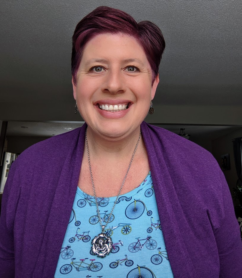 Melissa Wenzel light skin, short dark brown hair, brown eyes, large smile. Wearing a blue shirt with bicycle images printed and a purple sweater.