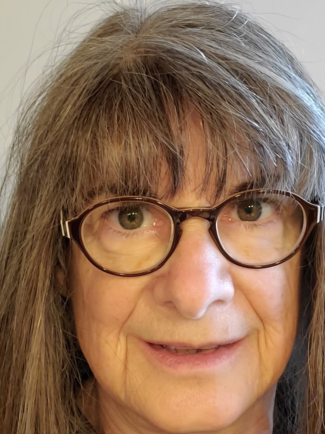 Laura Millberg Caucasian with brown/gray shoulder-length hair and bangs to eyebrows. Brown eyes and dark rimmed glasses. 