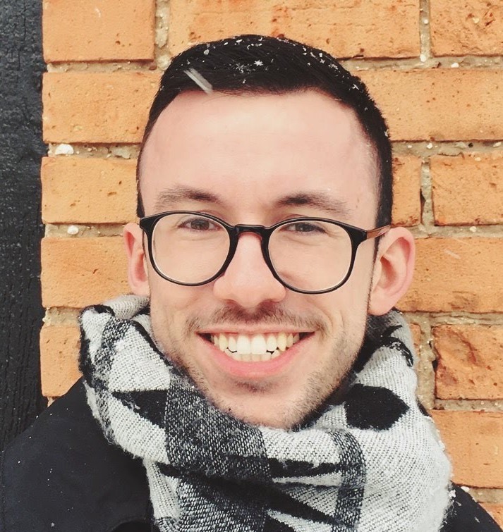 Cole Norgaarden white male with short dark hair, black rim round glasses, a winter scarf, and black jacket outside. 