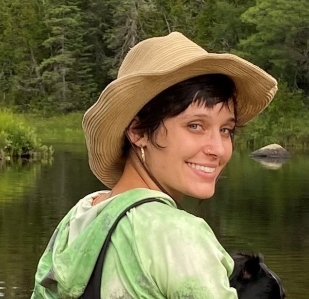 Rachel Morice - woman with short dark hair and light colored skin on a lake with a green blouse and black overalls wearing a straw hat