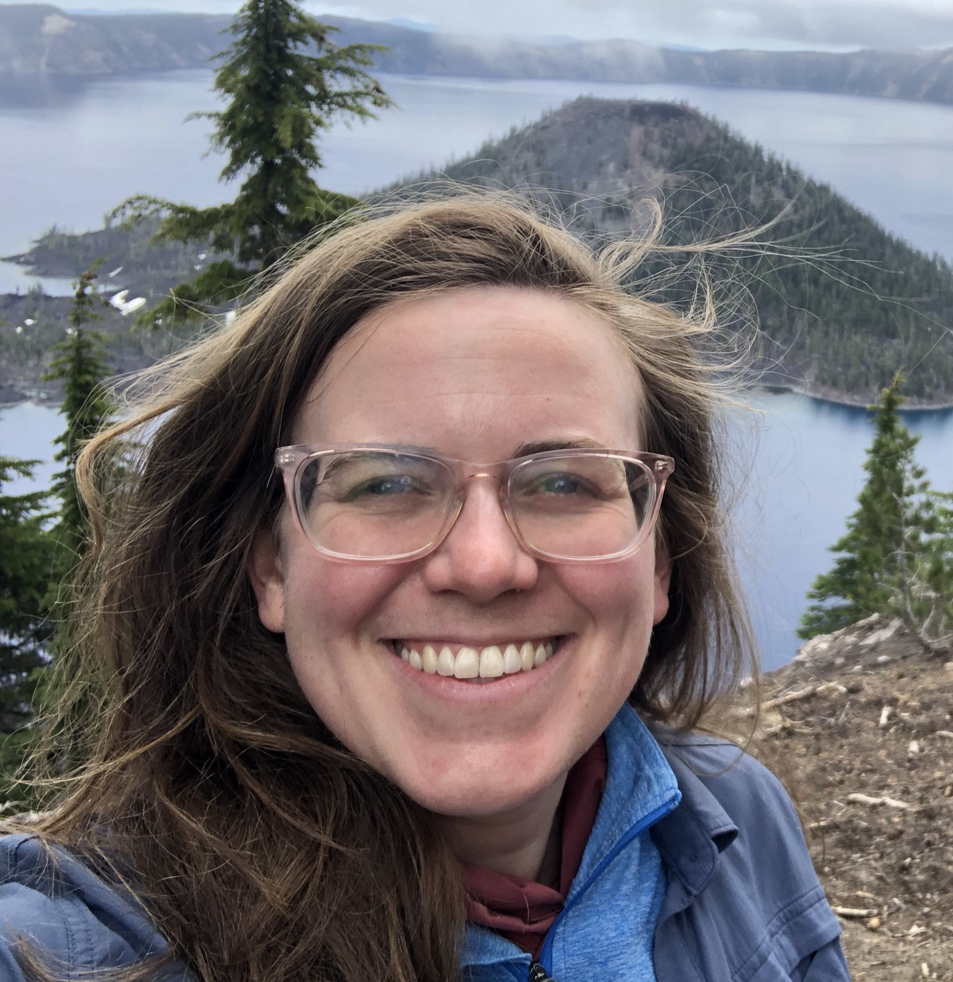 Headshot of Anna Callahan: long, straight light brown hair, glasses, smile, wearing a blue jacket standing in a forested area with a lake and island in the background