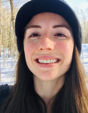 Amanda Wold - white woman smiling with below shoulder-length medium brown hair, wearing dark colored hat and jacket, standing outdoors with snow and trees in the background