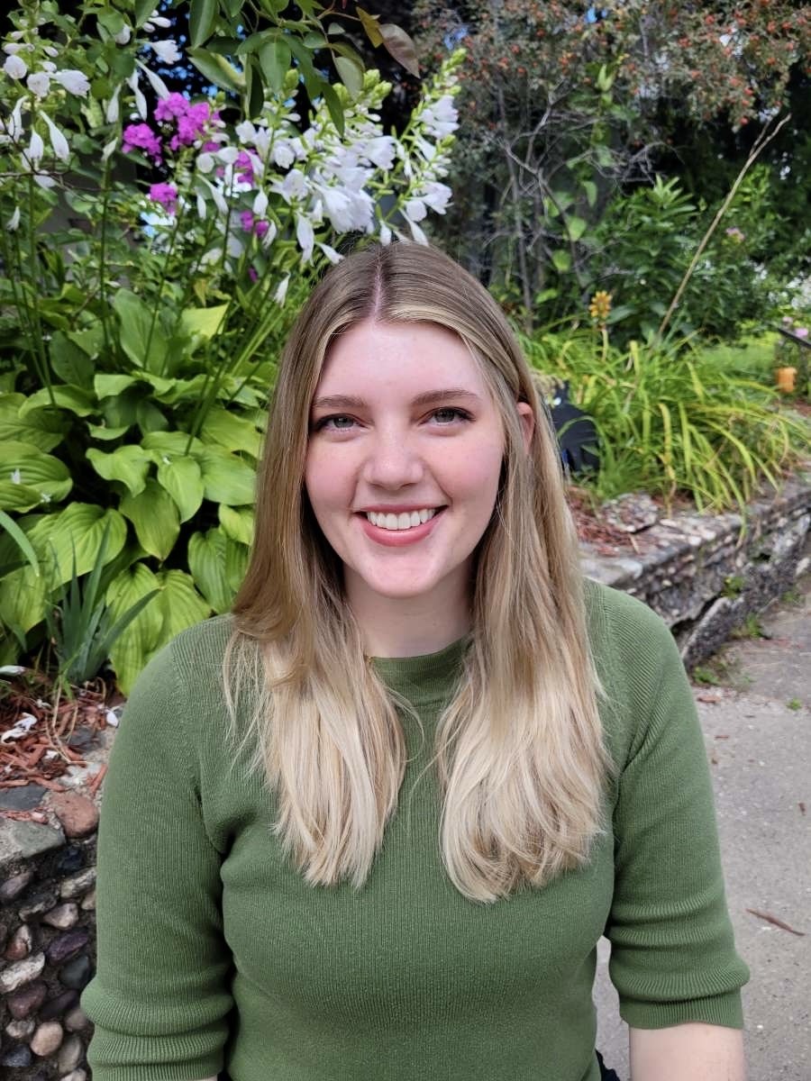 Lydia Godfrey headshot - blond female with below shoulder hair wearing a green mid-sleeve shirt, outside in front of a wall of greenery and floral