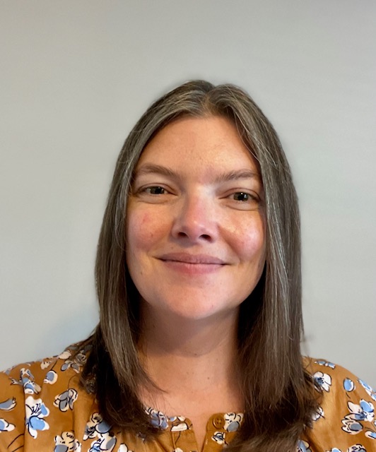 Nicole Smiley with an autumn orange floral blouse, brown hair with grey just past shoulder length, dark colored eyes, white skin, and a slight smile